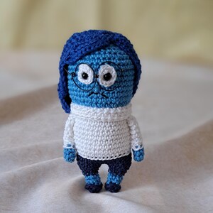 Könnte beinhalten: Eine blaue gehäkelte Figur mit einem weißen Pullover und blauen Hosen. Die Figur hat einen traurigen Ausdruck und trägt eine Brille.