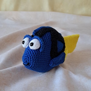 Könnte beinhalten: Ein gehäkelter blauer und schwarzer Fisch mit einer gelben Schwanzflosse. Der Fisch hat weiße Augen mit schwarzen Pupillen und ein kleines Lächeln.