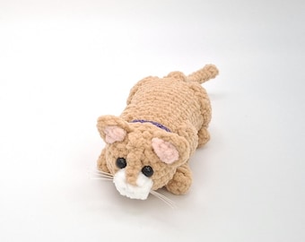 May include: A handmade, tan crochet cat toy with a purple collar, pink inner ears, and white muzzle. The cat has black eyes and long white whiskers. The toy is lying down on a white surface.