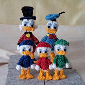 Puede incluir: Cinco patos de peluche tejidos a crochet, cada uno con un atuendo de color diferente. Los patos son todos blancos con ojos negros y picos naranjas. Los atuendos incluyen un traje rojo y azul, un atuendo de marinero azul y blanco, un suéter rojo, un suéter verde y un atuendo de marinero azul y blanco con una boina azul.
