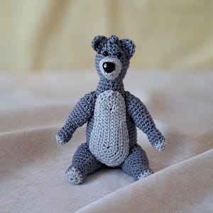 May include: A small gray crocheted bear with a black nose and black eyes. The bear is sitting on a white surface.