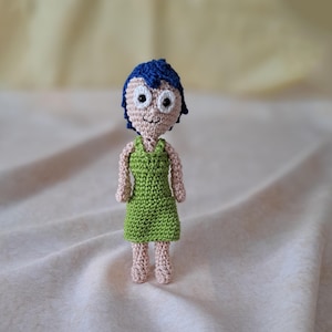 Könnte beinhalten: Eine gehäkelte Puppe mit blauen Haaren, einem grünen Kleid und weißen Augen. Die Puppe steht auf einer weißen Stoffoberfläche.