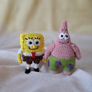 Könnte beinhalten: Gehäkelte Plüschtiere von SpongeBob Schwammkopf und Patrick Star. SpongeBob ist gelb mit einem braunen Gürtel und weißen Augen. Patrick ist rosa mit einer grünen Hose und lila Seestern-Designs.