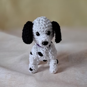 Puede incluir: Un perro dálmata de crochet blanco con manchas negras y orejas negras. El perro lleva un collar morado.