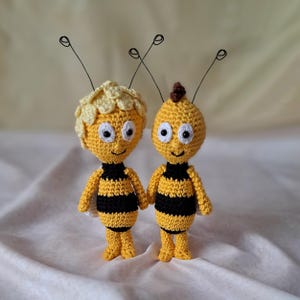 May include: Two crocheted bee plushies, one with a yellow head and the other with a brown head. Both have black and yellow striped bodies and white eyes.