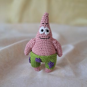 Könnte beinhalten: Eine rosa gehäkelte SpongeBob Schwammkopf Figur mit einem grün-violetten gehäkelten Outfit. Die Figur hat ein lächelndes Gesicht mit schwarzen Augen und einem weißen Umriss.