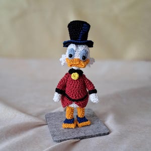 Puede incluir: Un juguete de peluche de ganchillo de un pato que lleva una chaqueta roja, pantalones negros, un sombrero de copa azul y negro y zapatos amarillos. El pato tiene la cabeza blanca y el pico amarillo.