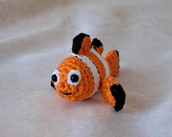 May include: A crocheted orange and white clownfish with black eyes and fins. The fish is lying on a white surface.