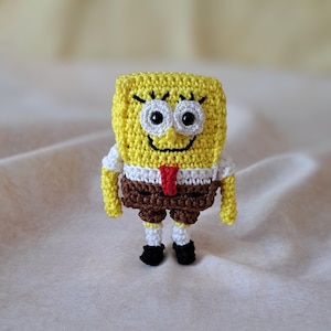 Könnte beinhalten: Eine gehäkelte Spongebob Schwammkopf Puppe mit einem gelben Körper, braunen Hosen, einem weißen Hemd und einer roten Krawatte. Die Puppe hat ein lächelndes Gesicht mit weißen Augen und einer schwarzen Nase.