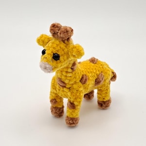 Könnte beinhalten: Ein kleines, handgefertigtes gelbes Häkel-Giraffen-Plüschtier mit braunen Flecken und Akzenten. Die Giraffe hat schwarze Augen und eine hellbraune Schnauze. Das Stofftier steht auf einer weißen Oberfläche.