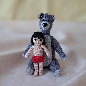 May include: A crocheted gray bear and a crocheted tan-skinned boy with black hair and red shorts. The bear is sitting with its arms outstretched and the boy is standing in front of the bear.