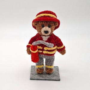 Könnte beinhalten: Ein handgefertigter brauner Teddybär, der als Feuerwehrmann gekleidet ist und eine rote Jacke mit gelben Streifen, eine graue Hose und einen roten Hut trägt. Der Bär hält einen roten Feuerlöscher. Der Bär steht auf einer grauen quadratischen Basis.