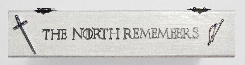 K&ouml;nnte beinhalten: Eine wei&szlig;e Holzkiste mit den Worten "The North Remembers" auf der Vorderseite eingraviert. Die Kiste hat ein silbernes Schwert und einen silbernen Pfeil mit einem blauen Edelstein an jedem Ende.