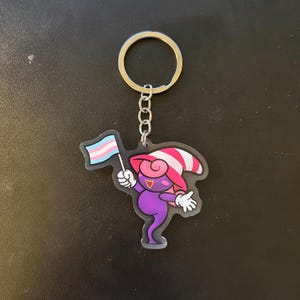 May include: A keychain featuring a cartoon character holding a transgender pride flag. The character is purple with a pink and white striped hat. The keychain has a silver ring and chain.