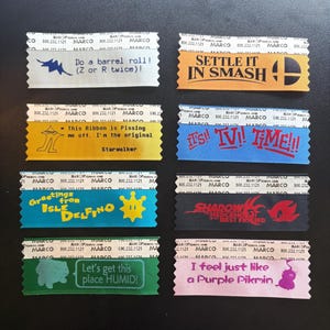 May include: Eight rectangular fabric ribbons with serrated edges, each featuring unique printed text and graphics. Designs include references to video games like Smash, Starwalker, Isle Delfino, Shadow the Hedgehog, Bulbasaur, and Pikmin.