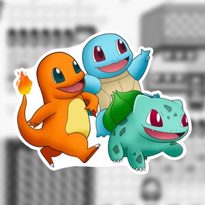 May include: A sticker featuring three cartoon characters: an orange Charmander with a flame, a blue Squirtle, and a green Bulbasaur. The characters have large eyes and are smiling. The background is blurred.