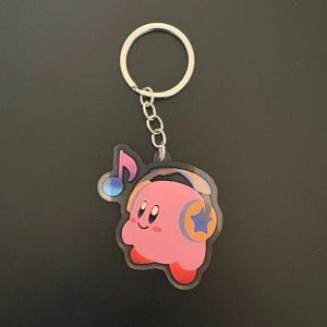 May include: A pink Kirby keychain with a silver keyring and chain. The keychain features Kirby wearing headphones with a star on them and a musical note. The background is a solid black color.