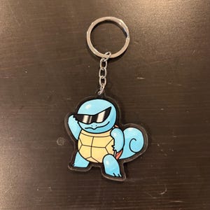 May include: A blue keychain featuring a cartoon turtle wearing sunglasses. The turtle has a yellow shell and is holding a pose. The keychain has a silver metal ring and chain.