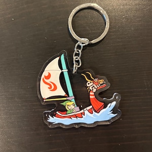 May include: A clear acrylic keychain featuring a cartoon boat with a white sail and red accents. The boat is sailing on blue waves, and a cartoon character is in the boat. The keychain has a silver metal ring.