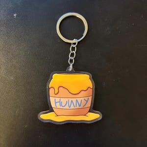 May include: A keychain featuring a cartoon honey pot design. The pot is orange with a yellow honey spill and the word "HUNNY" in blue. The keychain has a silver metal ring and chain.