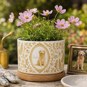 May include: A white ceramic planter with a gold botanical pattern and a central oval frame containing an illustration of a golden retriever and the name Charlie. The planter sits on a round wooden base and holds pink cosmos flowers.