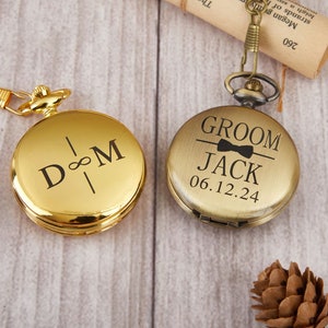 May include: Two gold pocket watches with engraved designs. The watch on the left has the initials "D & M" with an infinity symbol between them. The watch on the right has the word "Groom" above the name "Jack" and the date "06.12.24".