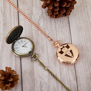 Puede incluir: Dos relojes de bolsillo con cadenas. Un reloj tiene una esfera blanca con marcadores de hora romanos negros y un acabado dorado. El otro reloj tiene un acabado de oro rosa y un diseño grabado de un esmoquin con la palabra "Gift" en él.