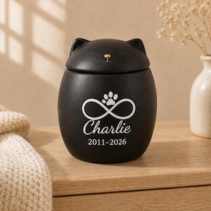 May include: A matte black ceramic pet urn shaped like a cat head with a gold-coloured nose. The front features a white infinity symbol with a paw print, the name Charlie, and the years 2011-2026 engraved in a clean, white, sans-serif font.