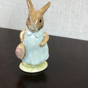 May include: A small porcelain figurine of a brown rabbit wearing a blue dress and holding a pink basket. The rabbit has brown eyes and a white nose.