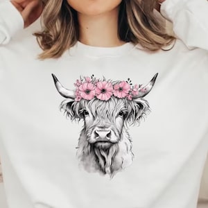 May include: White sweatshirt featuring a detailed illustration of a Highland cow with a floral crown. The cow is depicted in black and white, with pink flowers adorning its head. The sweatshirt has long sleeves and a crew neck.