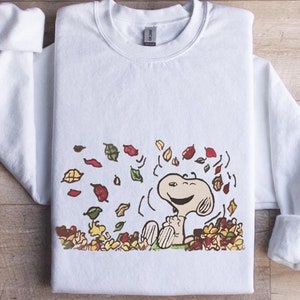 May include: White sweatshirt featuring a cartoon Snoopy character surrounded by colorful autumn leaves. The design includes Snoopy sitting in a pile of leaves. The sweatshirt has a crew neck and long sleeves.