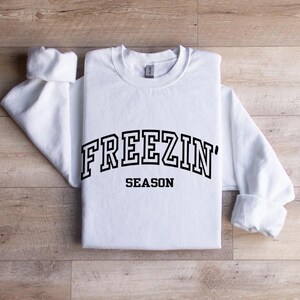 Może przedstawiać: Biała bluza z długim rękawem z czarnym napisem "FREEZIN' SEASON" w stylu uniwersyteckim.