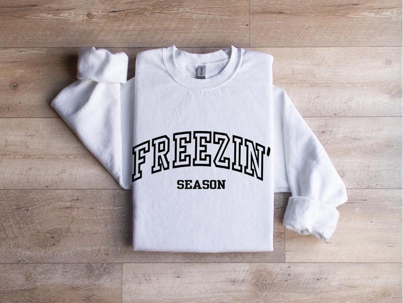 Może przedstawiać: Biała bluza z długim rękawem z czarnym napisem "FREEZIN' SEASON" w stylu uniwersyteckim.