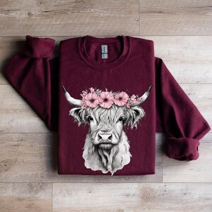 May include: A maroon sweatshirt featuring a detailed illustration of a Highland cow. The cow has a white face, long fur, and is adorned with a floral crown of pink flowers. The sleeves are cuffed, and the background is a wooden surface.