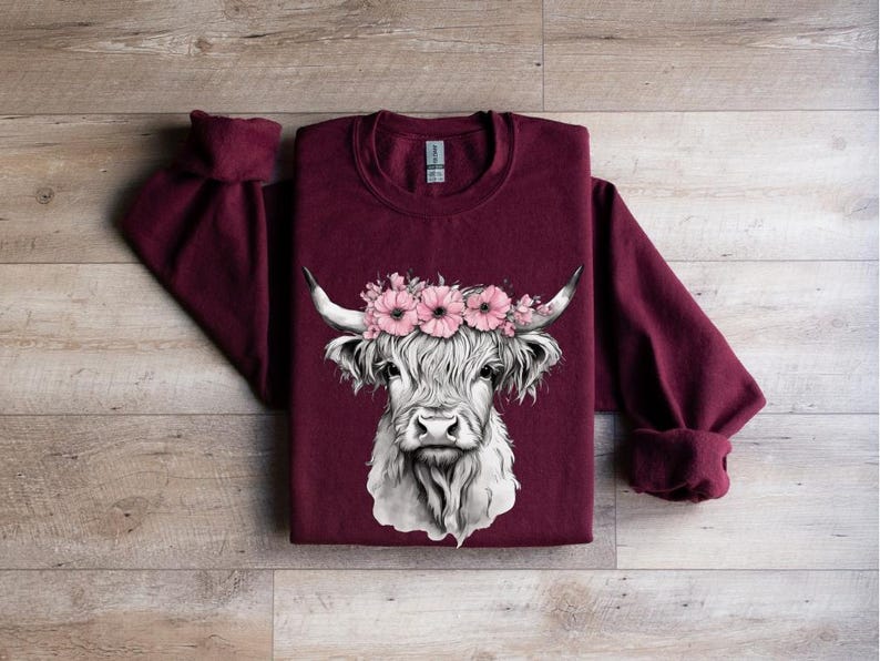 May include: A maroon sweatshirt featuring a detailed illustration of a Highland cow. The cow has a white face, long fur, and is adorned with a floral crown of pink flowers. The sleeves are cuffed, and the background is a wooden surface.