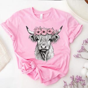 May include: A pink t-shirt featuring a detailed black and white illustration of a Highland cow wearing a floral crown. The cow has long, shaggy fur and curved horns. The shirt is made of soft material and has rolled-up sleeves.