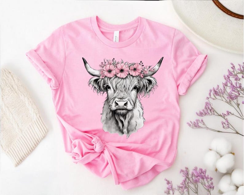 May include: A pink t-shirt featuring a detailed black and white illustration of a Highland cow wearing a floral crown. The cow has long, shaggy fur and curved horns. The shirt is made of soft material and has rolled-up sleeves.