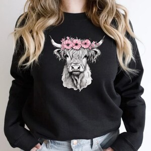 May include: Black sweatshirt featuring a detailed grayscale illustration of a Highland cow's head. The cow is adorned with a crown of pink flowers. The design is centered on the front of the long-sleeved top.