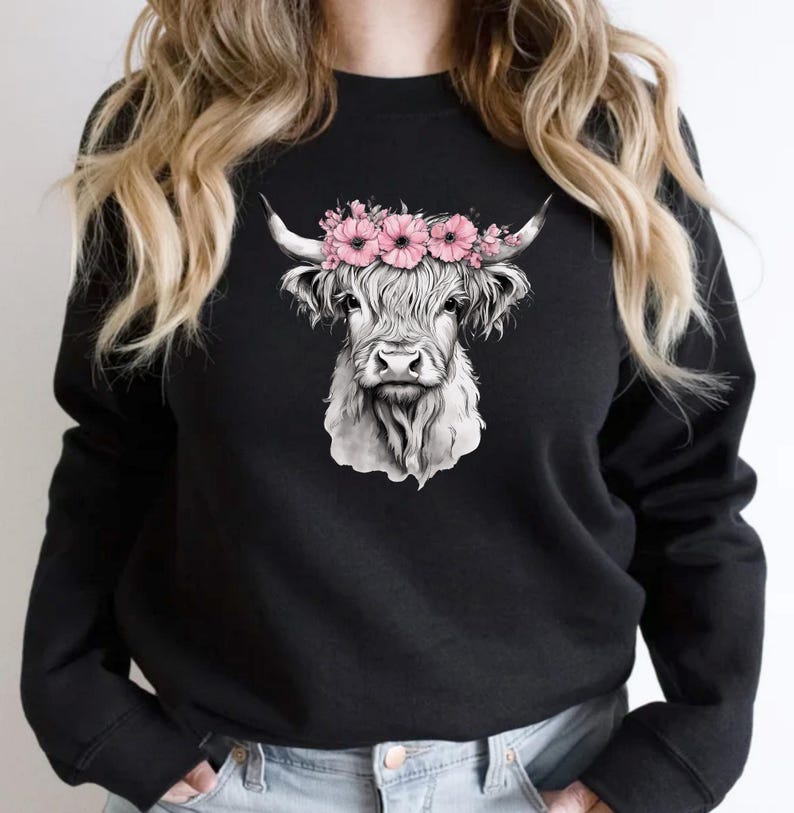 May include: Black sweatshirt featuring a detailed grayscale illustration of a Highland cow's head. The cow is adorned with a crown of pink flowers. The design is centered on the front of the long-sleeved top.