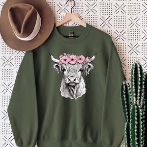 May include: An olive green sweatshirt featuring a detailed black and white illustration of a Highland cow wearing a pink flower crown. The sweatshirt is hanging on a wooden hanger. A brown hat is to the left of the sweatshirt.
