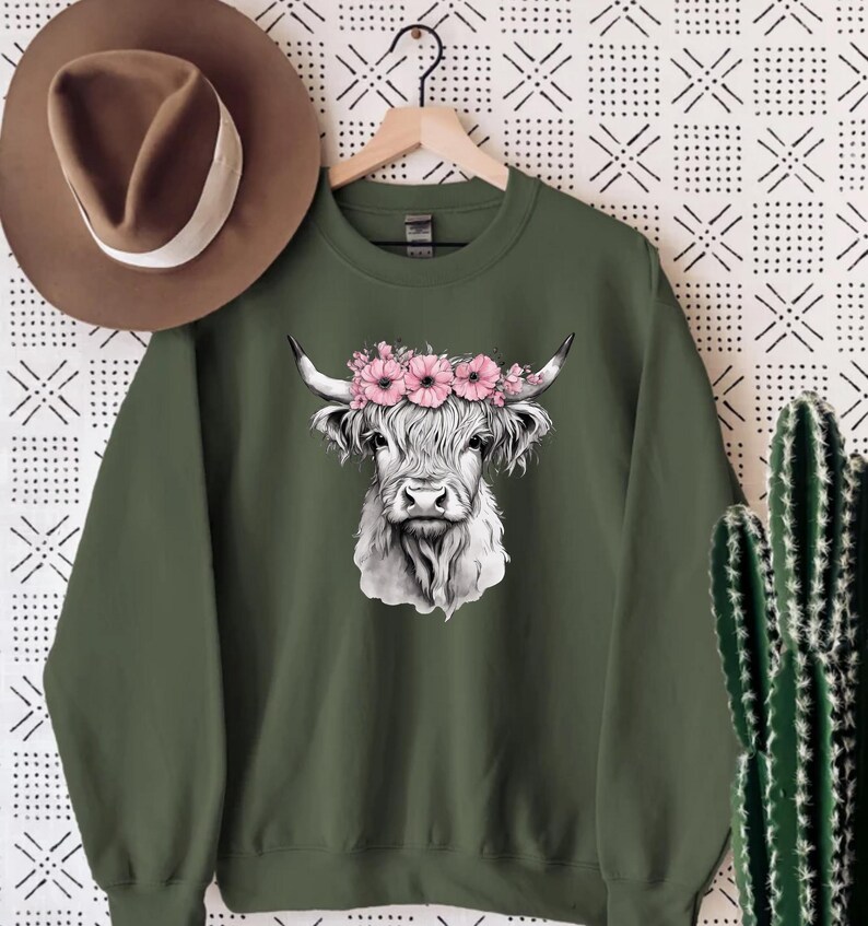 May include: An olive green sweatshirt featuring a detailed black and white illustration of a Highland cow wearing a pink flower crown. The sweatshirt is hanging on a wooden hanger. A brown hat is to the left of the sweatshirt.