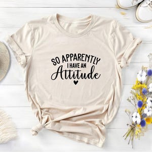 K&ouml;nnte beinhalten: Ein beiges T-Shirt mit dem Text "So Apparently I Have An Attitude" in schwarzer Schrift und einem kleinen schwarzen Herz.