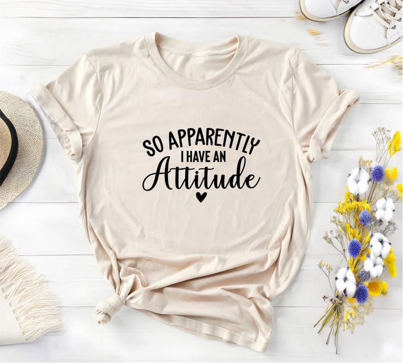 K&ouml;nnte beinhalten: Ein beiges T-Shirt mit dem Text "So Apparently I Have An Attitude" in schwarzer Schrift und einem kleinen schwarzen Herz.