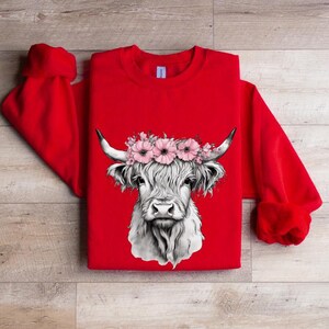 May include: A red sweatshirt featuring a detailed black and white illustration of a Highland cow's face. The cow is adorned with a floral crown of pink flowers. The sweatshirt has long sleeves and a crew neck.