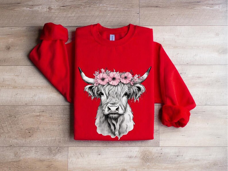 May include: A red sweatshirt featuring a detailed black and white illustration of a Highland cow's face. The cow is adorned with a floral crown of pink flowers. The sweatshirt has long sleeves and a crew neck.
