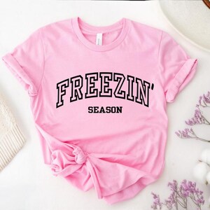 Może przedstawiać: R&oacute;żowa koszulka z czarnym napisem "FREEZIN' SEASON"