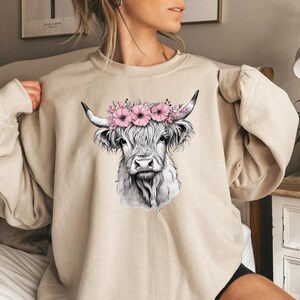 May include: Beige sweatshirt featuring a black and white illustration of a Highland cow wearing a pink floral crown. The cow has long, shaggy fur and curved horns. A comfortable and stylish top for casual wear.