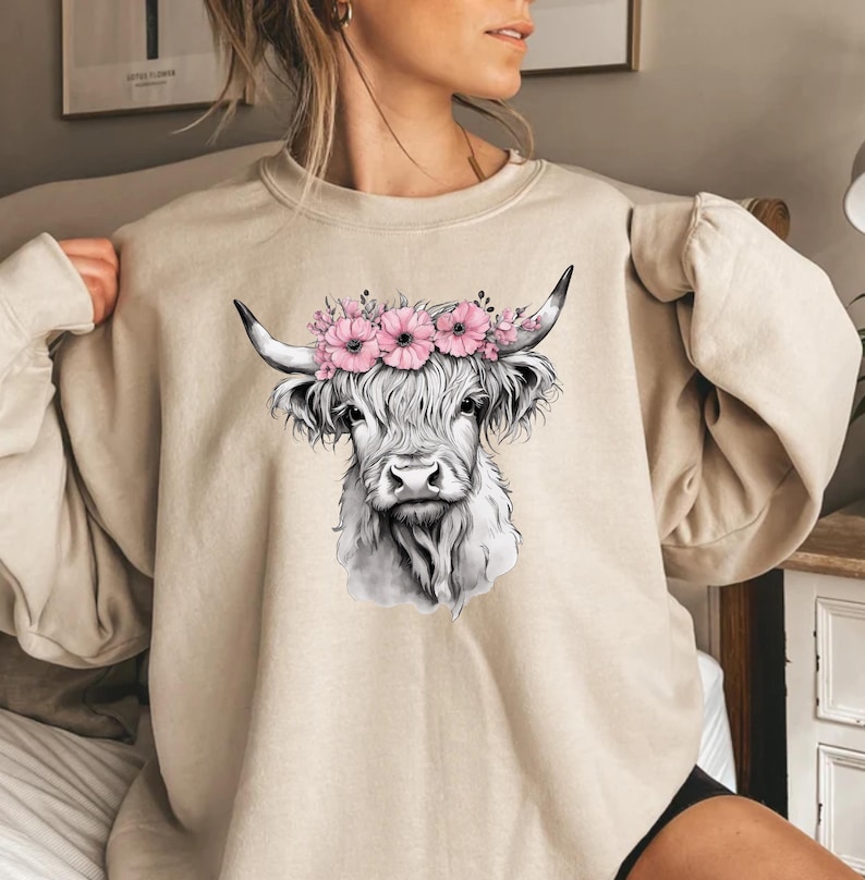 May include: Beige sweatshirt featuring a black and white illustration of a Highland cow wearing a pink floral crown. The cow has long, shaggy fur and curved horns. A comfortable and stylish top for casual wear.