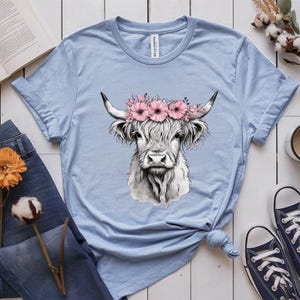 May include: Light blue t-shirt featuring a detailed illustration of a Highland cow wearing a floral crown. The cow is depicted in black and white, with long, shaggy fur and curved horns. The shirt is styled with rolled-up sleeves and a knotted hem.