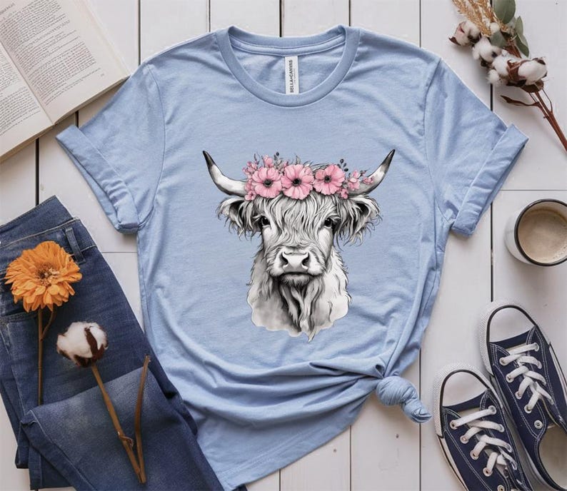 May include: Light blue t-shirt featuring a detailed illustration of a Highland cow wearing a floral crown. The cow is depicted in black and white, with long, shaggy fur and curved horns. The shirt is styled with rolled-up sleeves and a knotted hem.